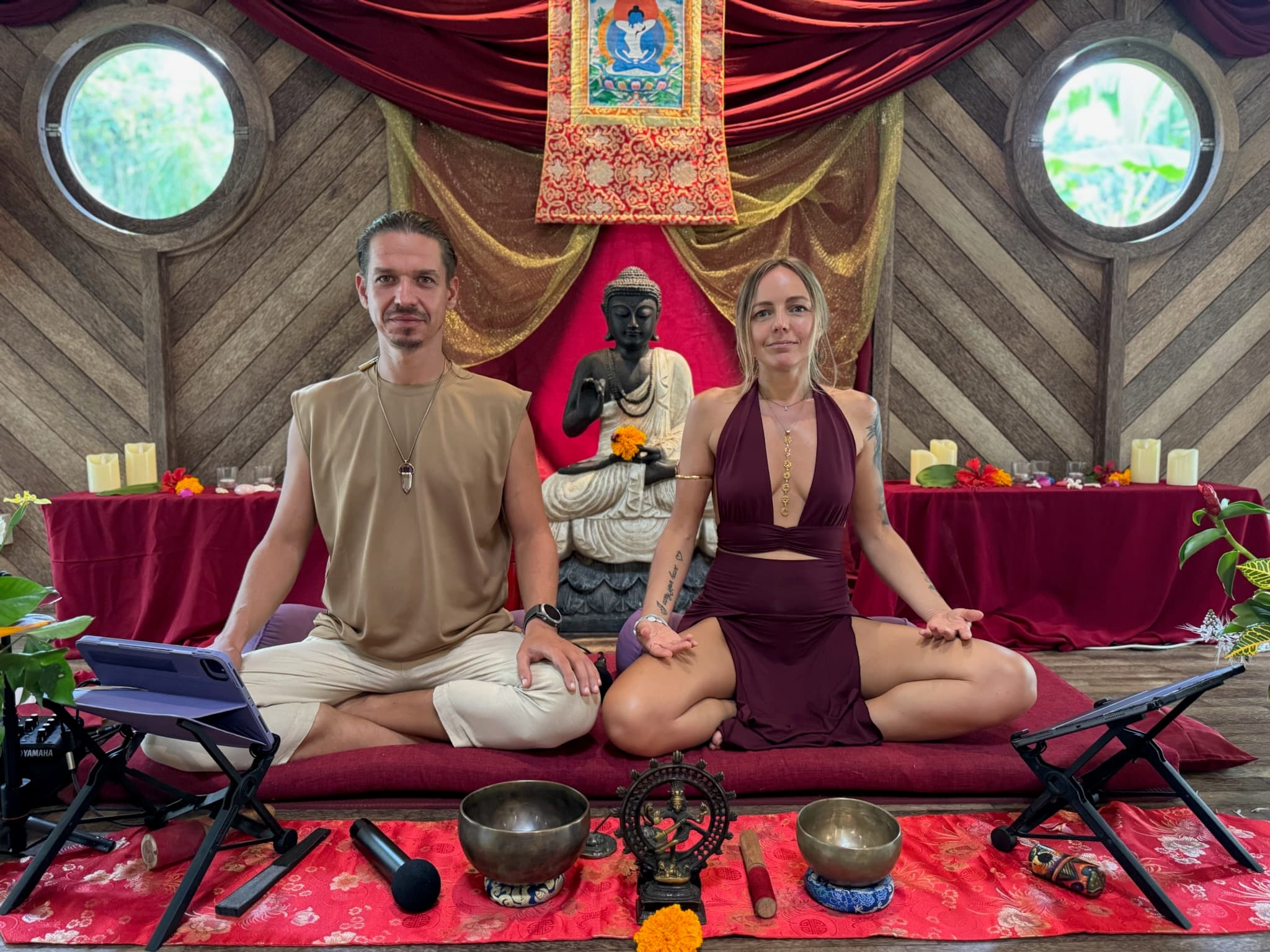 Lina and Ritam seated together in meditation posture on a burgundy mat in a wooden temple space, a stone Buddha, thangka, and singing bowls around them.