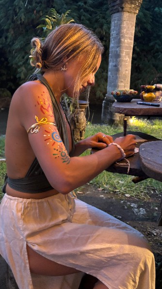 Lina at dusk with painted sacred symbols on her arm, preparing quietly at a wooden table in a garden.