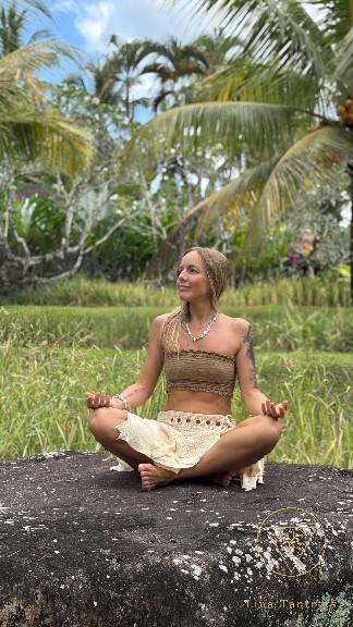 Lina seated cross-legged in meditation on a large stone, surrounded by tropical greenery and palms.