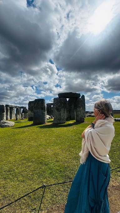 Lina in a cream shawl seen from behind, standing before the ancient standing stones of Stonehenge under a dramatic sky.