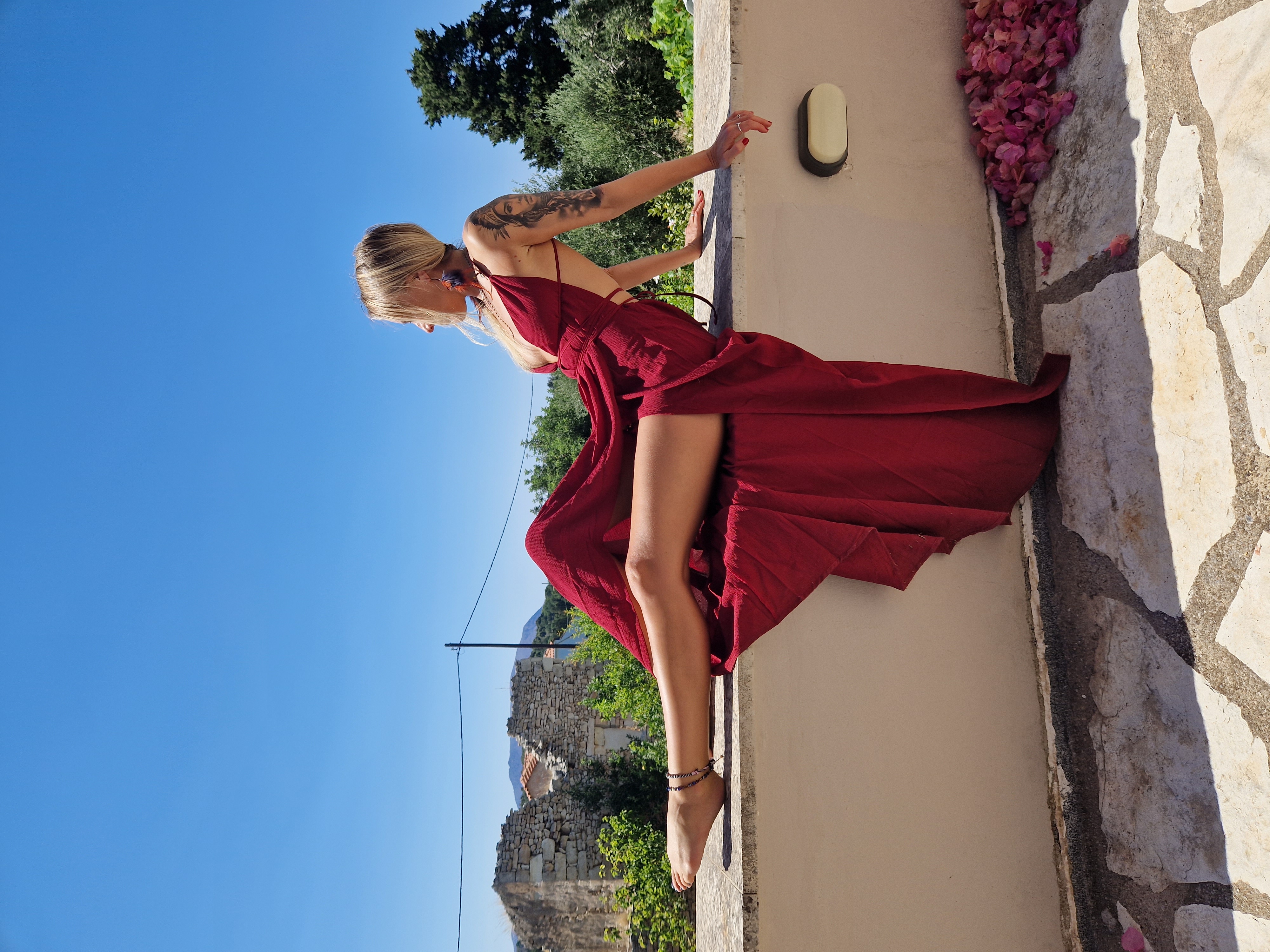 Lina reclining along a sunlit stone wall in a flowing wine-red dress against a clear blue sky, with rose petals scattered nearby.