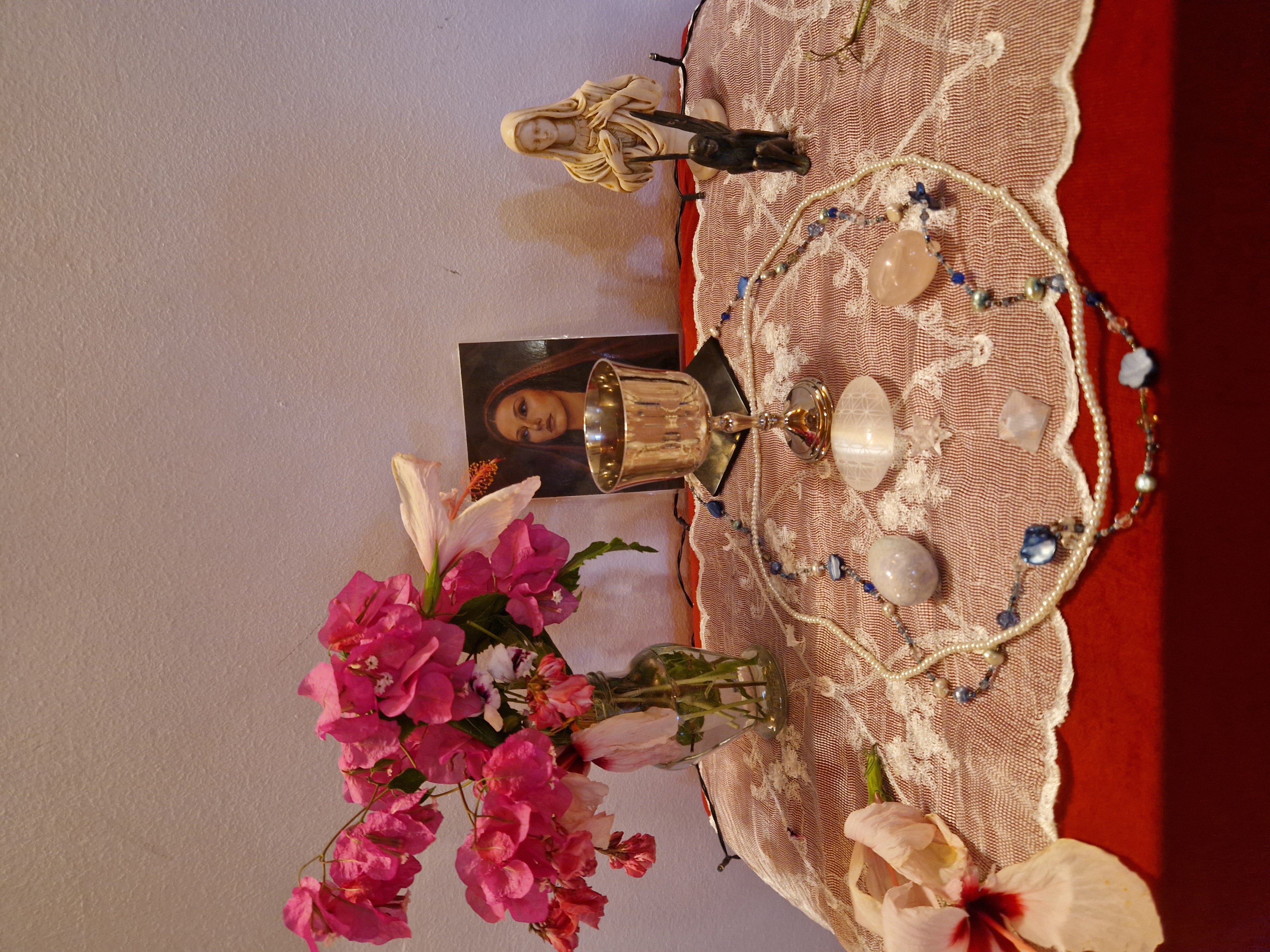 A devotional altar with a silver chalice, a rose-veiled image of the sacred feminine, a Madonna statue, crystals, pearls, and bougainvillea on lace.