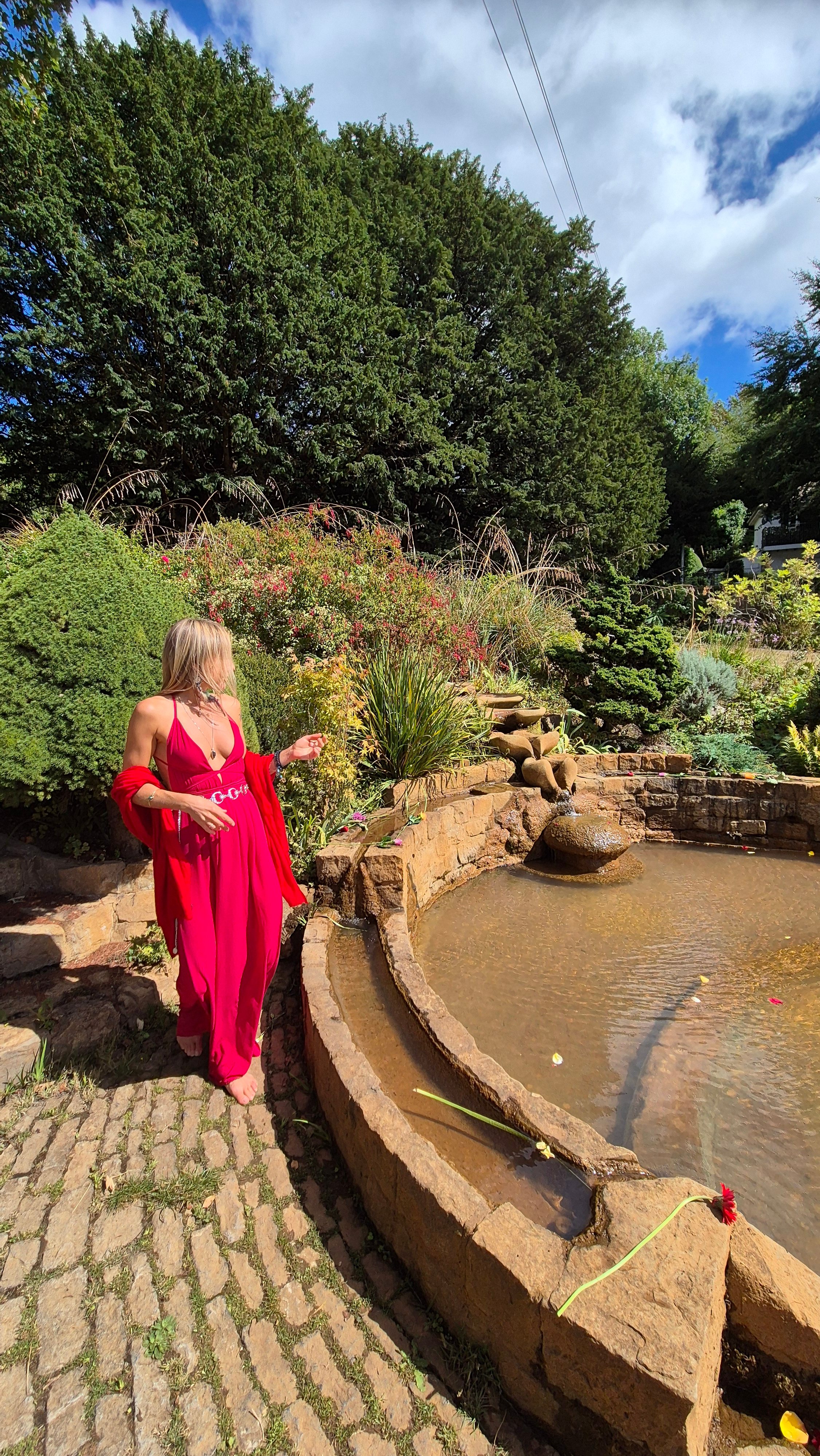 Lina in a flowing crimson dress standing beside a stone garden pool with a gentle fountain, surrounded by greenery.