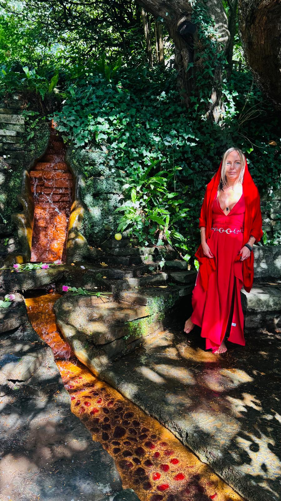 Lina in a red veil and flowing red gown standing beside a natural spring in a lush ivy garden, with rose petals in the water.