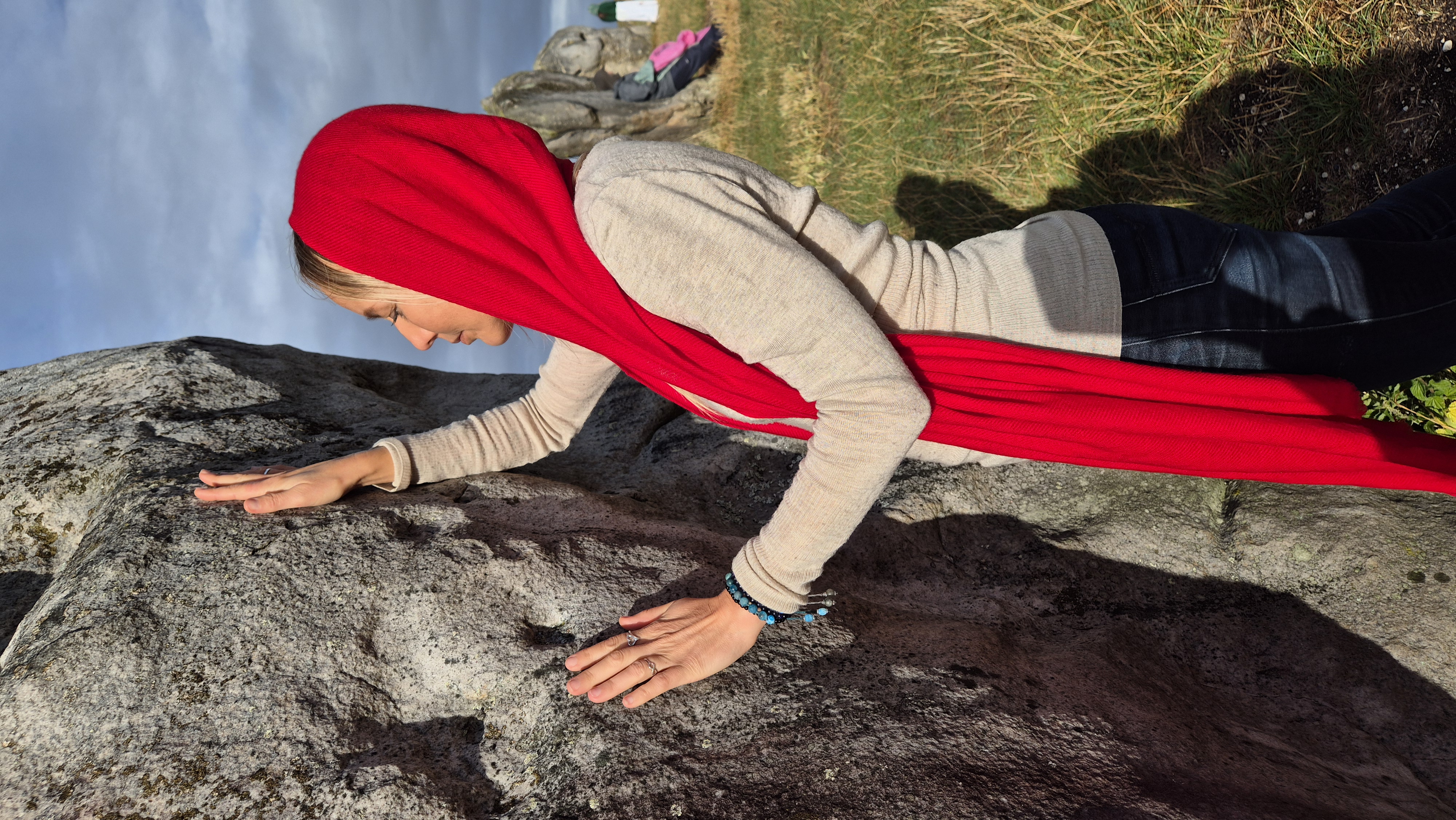 Lina in a red veil resting her hands in devotion against an ancient standing stone at a stone circle.