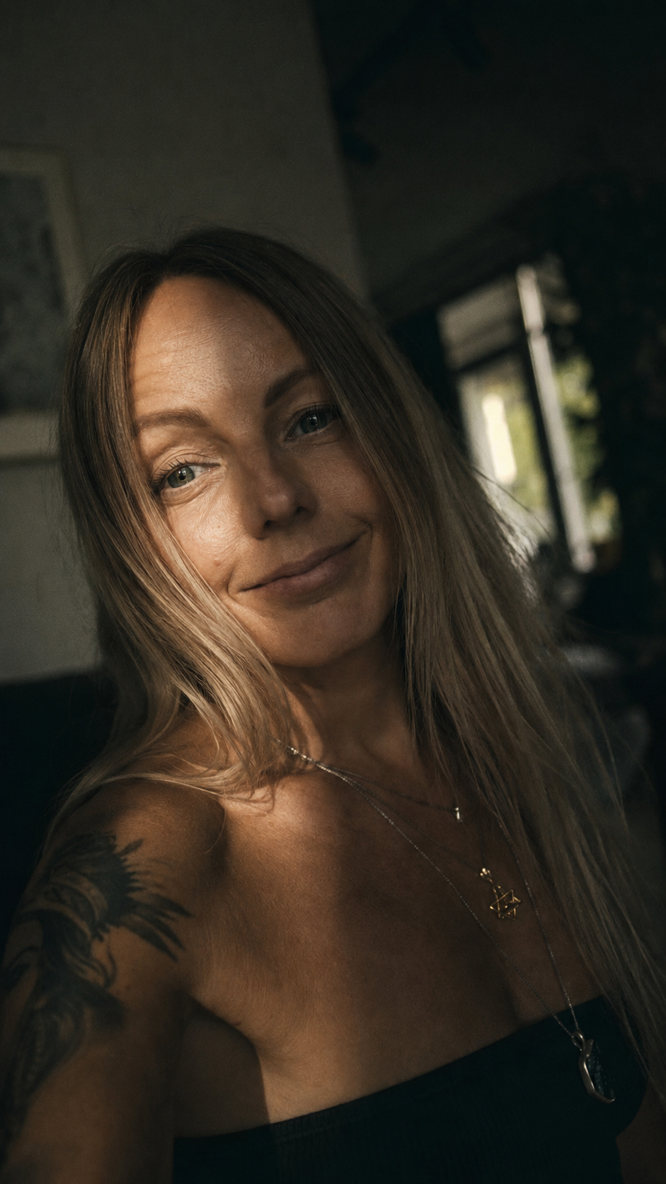 Lina smiling softly in a warmly lit room, with layered necklaces and an arm tattoo.